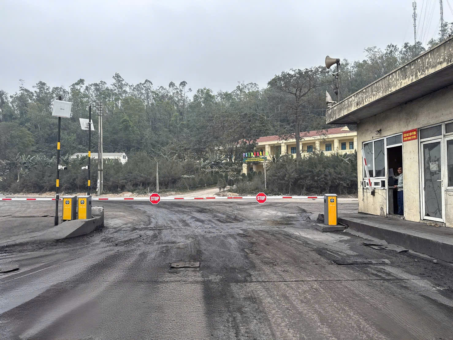 Trạm kiểm soát với barie và biển báo dừng trên con đường phủ bụi, phía sau là tòa nhà và hàng cây